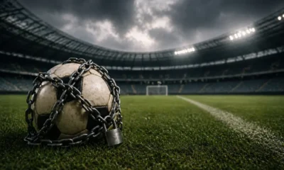 Un pallone incatenato con un lucchetto su un campo da calcio, sullo sfondo nubi nere